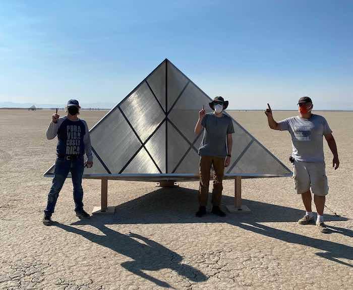 Three men holding up their index fingers to indicate one, standing in front of a 2.8 meter triangular metal corner reflector